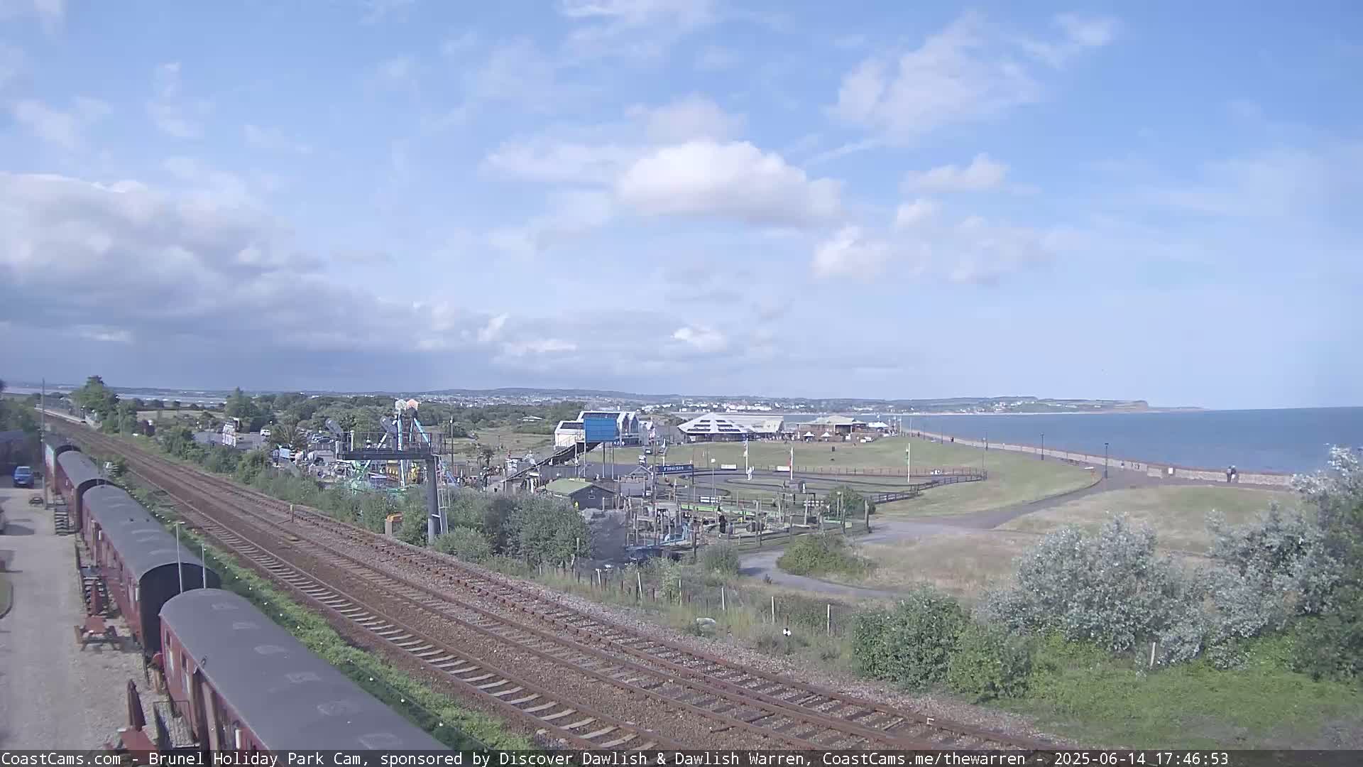 A seaside amusement park with a playground and train tracks is visible under a partly cloudy sky, next to a calm ocean.