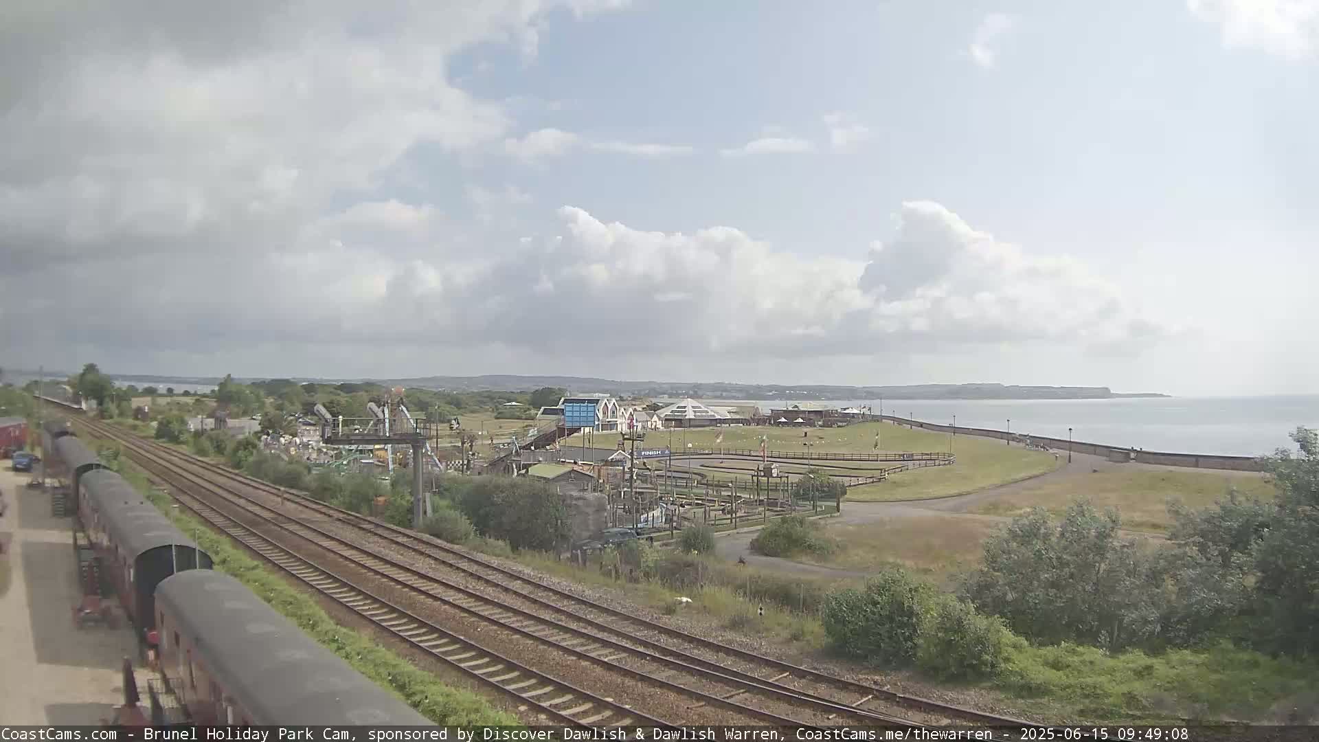 A seaside amusement park is visible near a train track, under a partly cloudy sky.