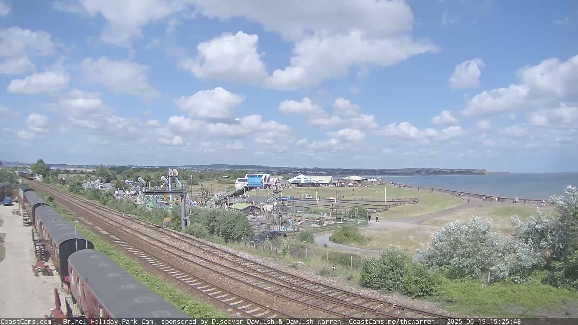 A seaside amusement park with a playground and train tracks is visible under a partly cloudy sky, next to a calm body of water with a shoreline where people are walking.