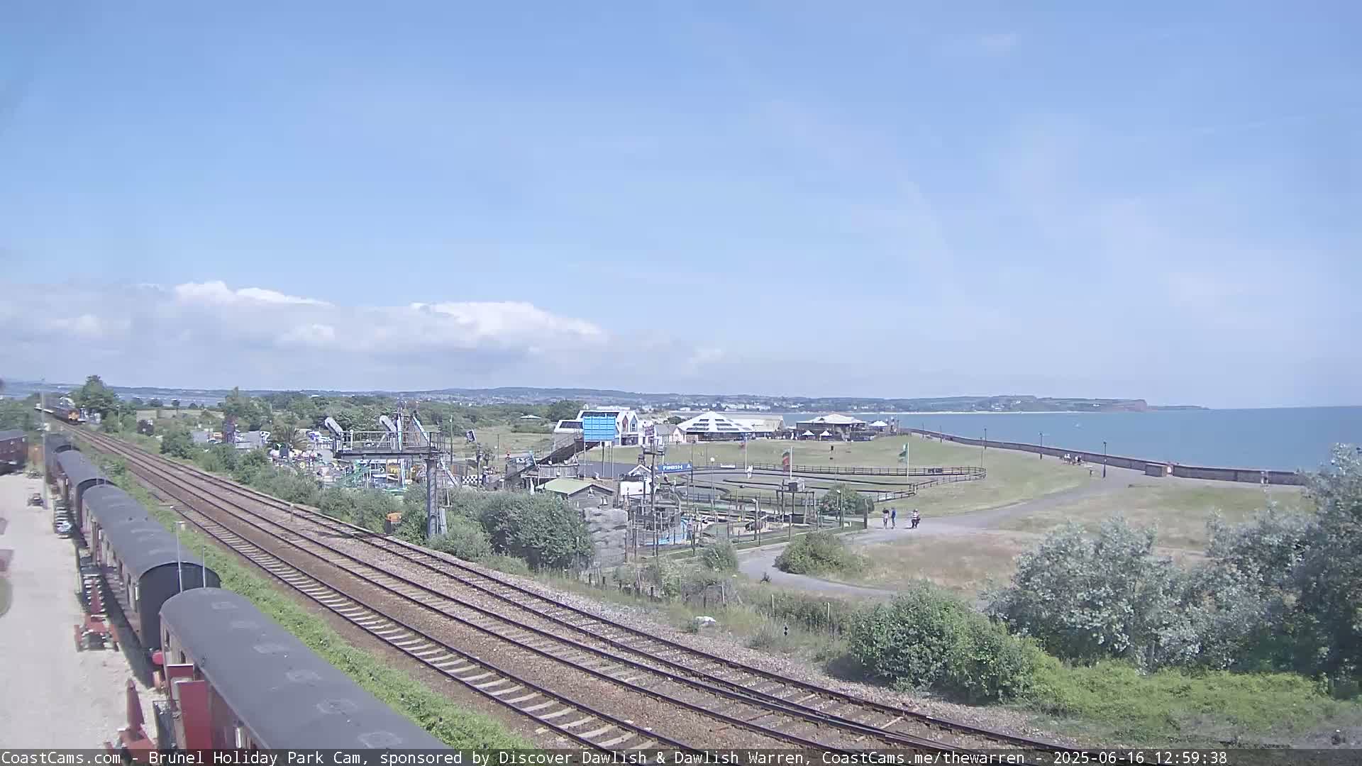 A sunny day reveals a seaside amusement park, train tracks, and railway cars near a calm ocean.