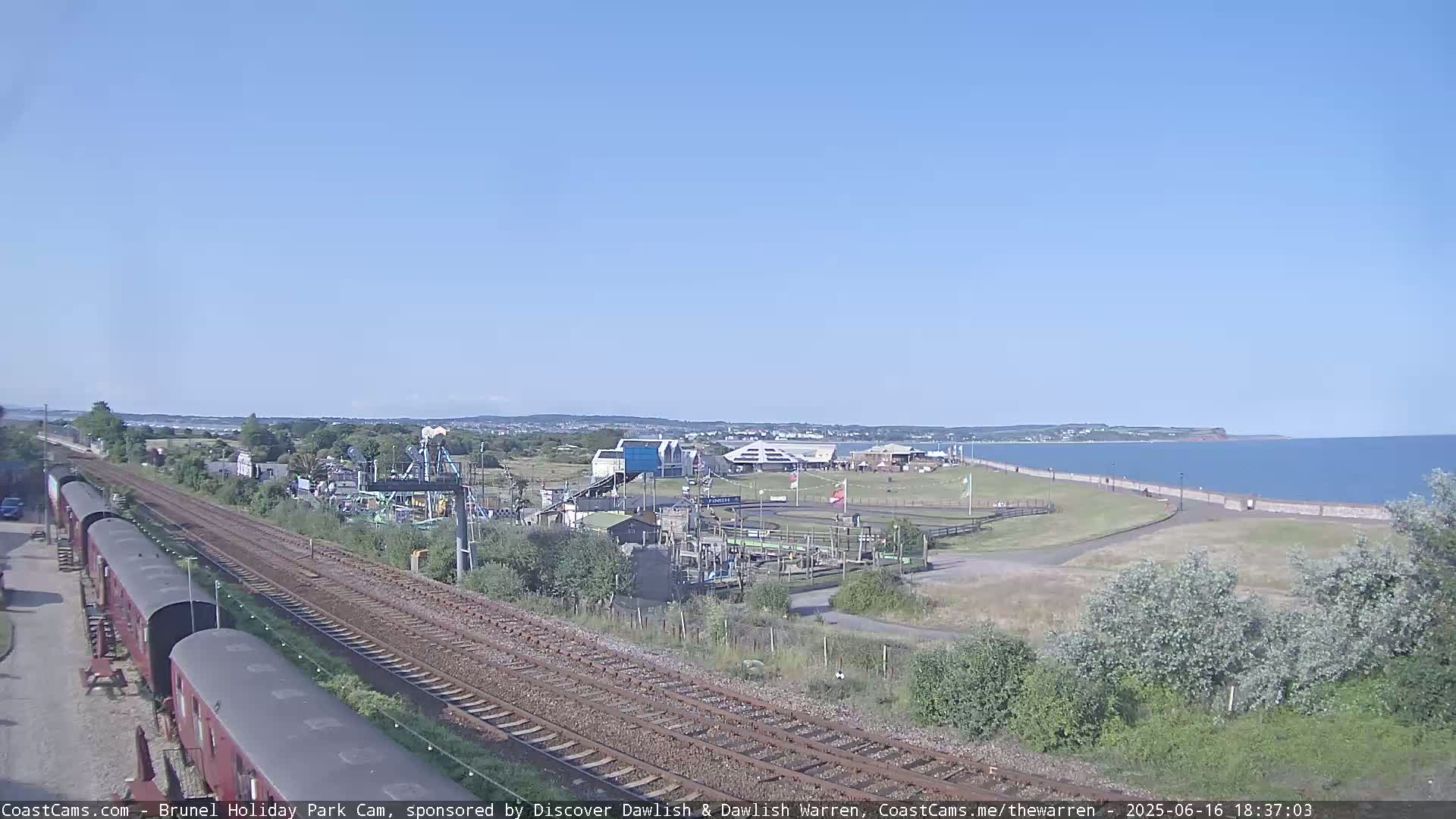 A clear sunny day shows a coastal scene with a train track in the foreground leading to a seaside park and a calm ocean beyond.