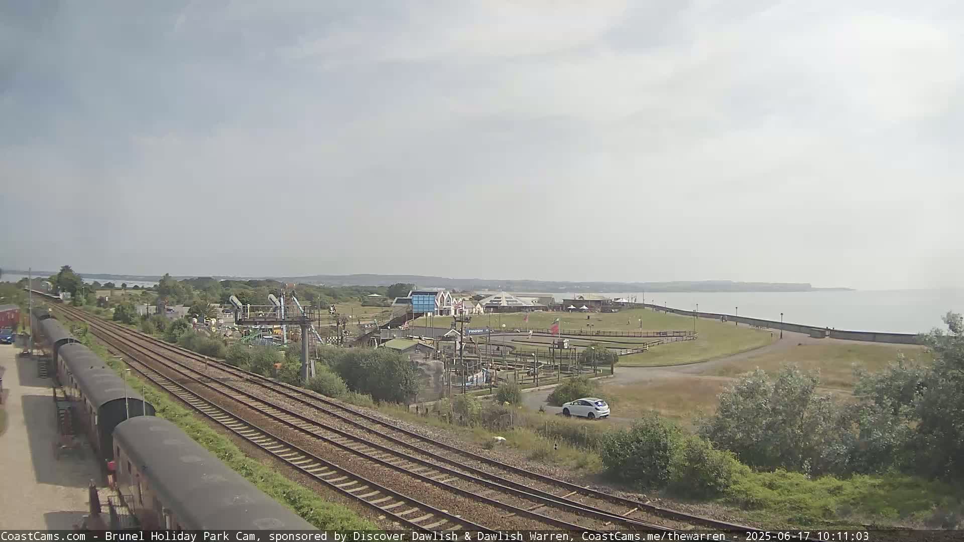 A seaside amusement park with a go-kart track and playground is visible near a body of water under an overcast sky, with railway tracks running alongside.