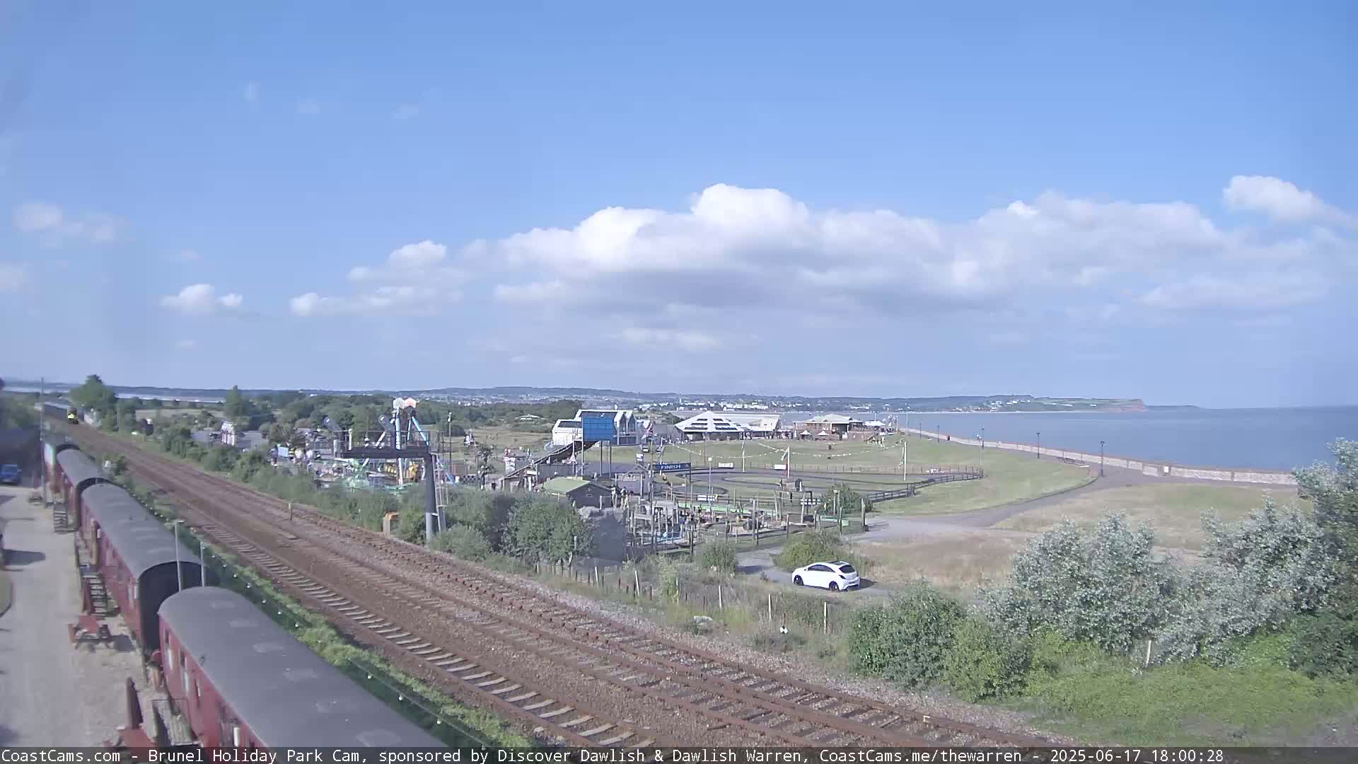 A partly cloudy day shows a seaside amusement park next to train tracks, with a calm body of water and a shoreline in the background.