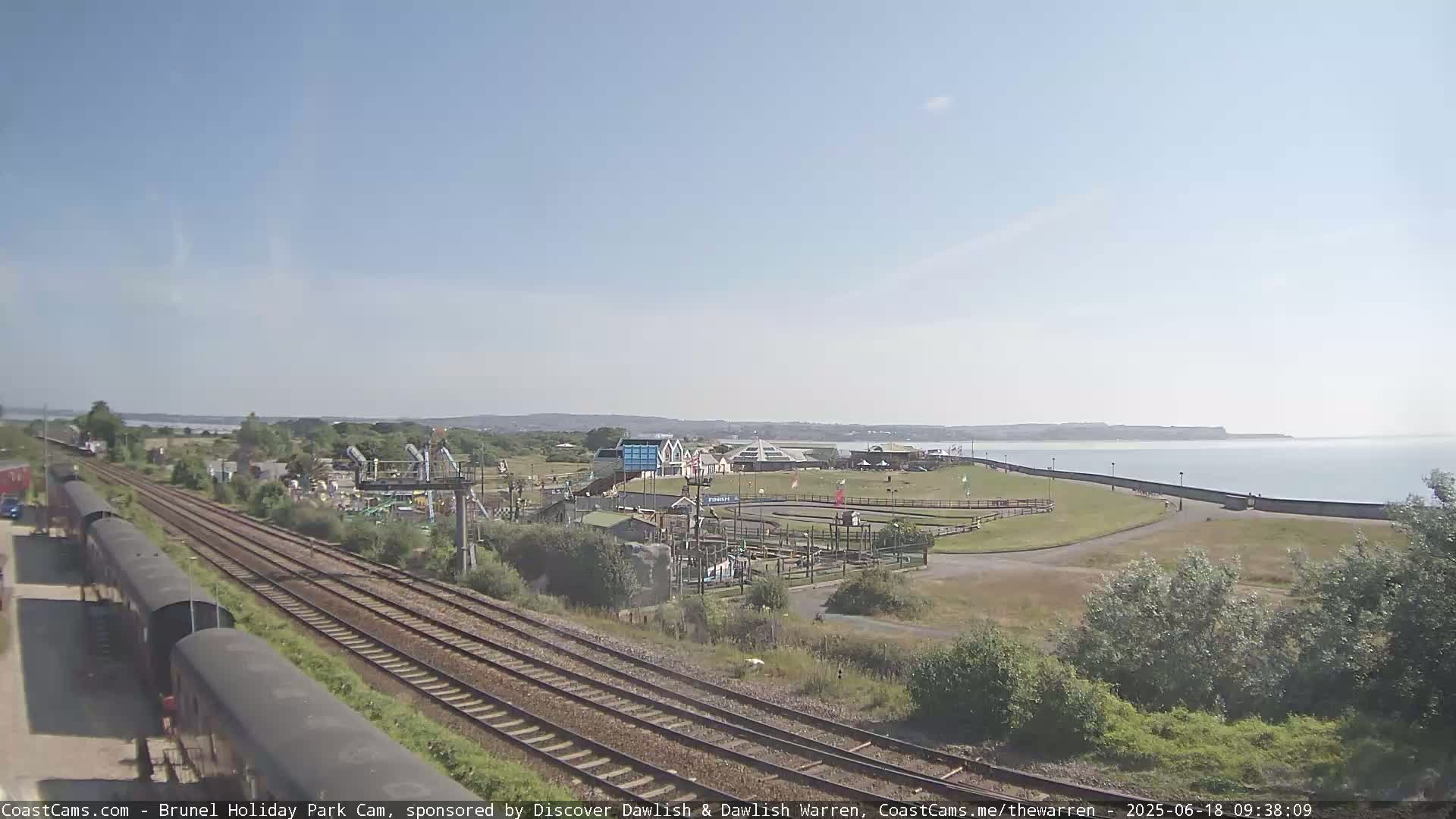A seaside amusement park with a go-kart track and playground is visible beside train tracks, under a clear blue sky.