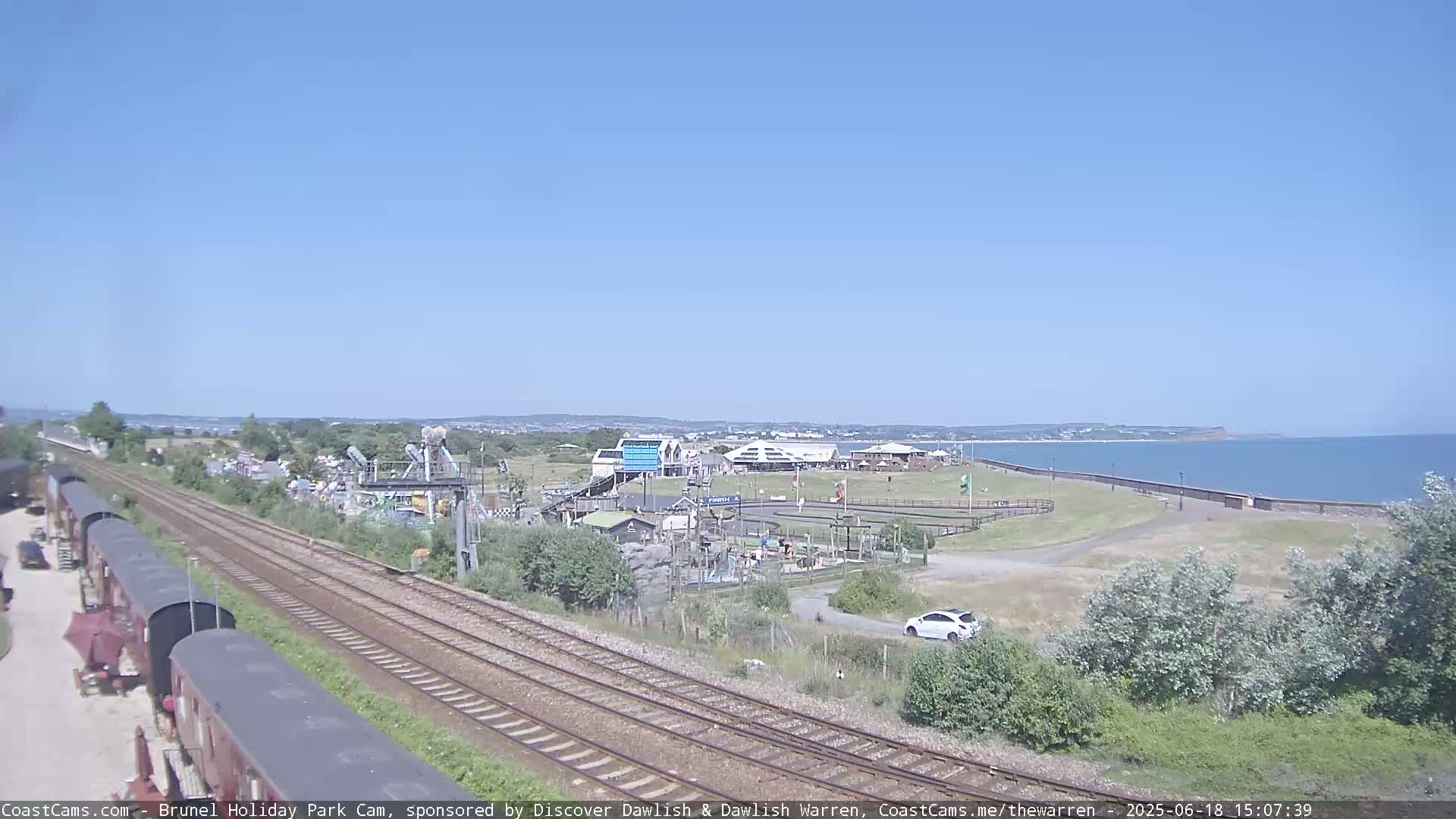 A seaside amusement park with train tracks and parked railway cars is visible under a clear blue sky.