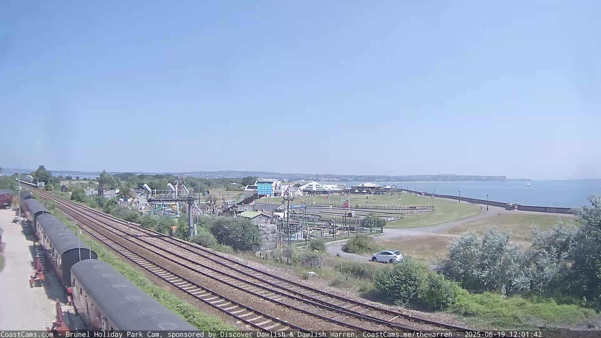 A sunny day overlooks a seaside amusement park with railway tracks in the foreground and a calm ocean in the background.