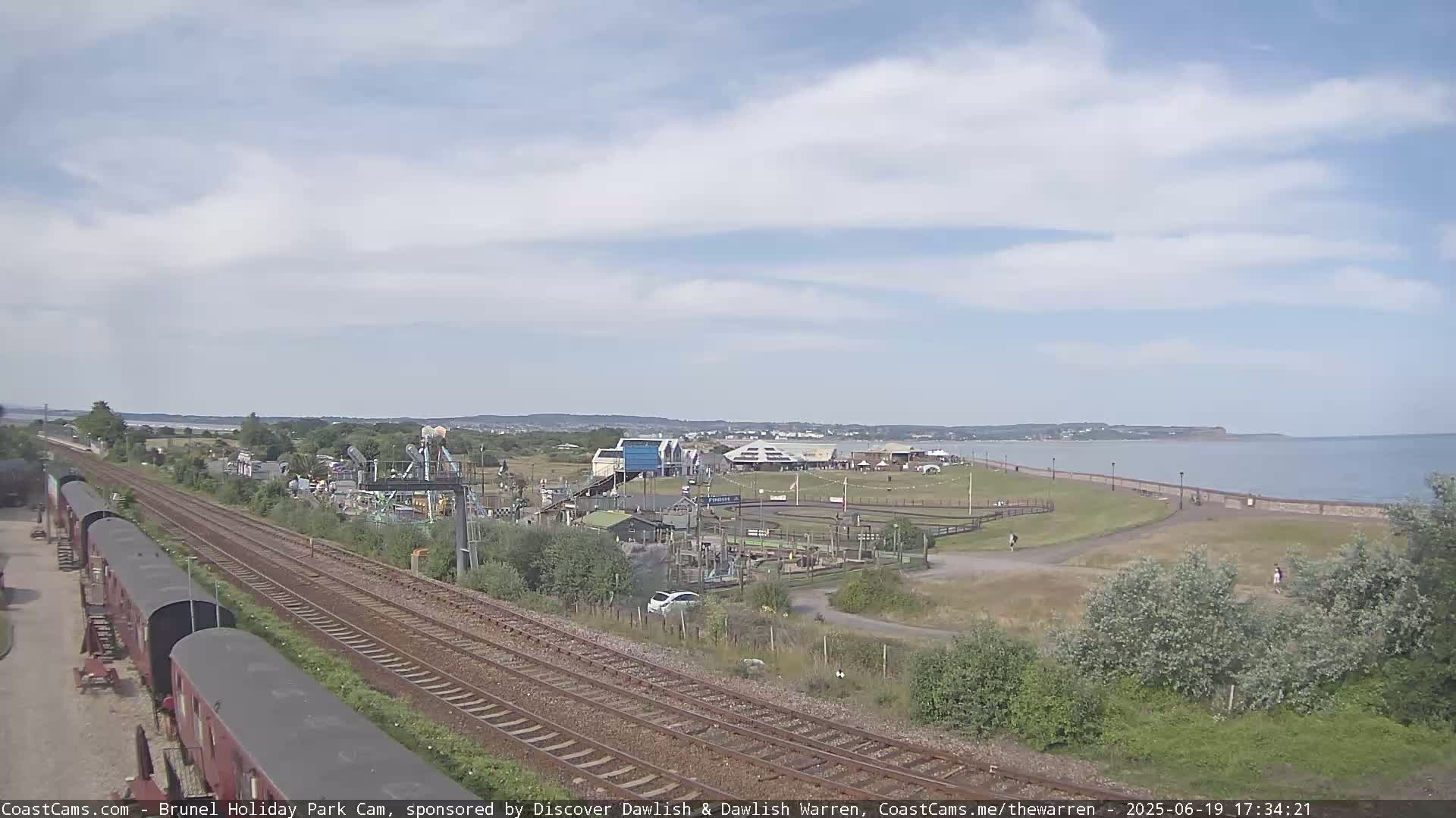 A seaside amusement park with a playground and train tracks is visible under a partly cloudy sky, next to a calm body of water.