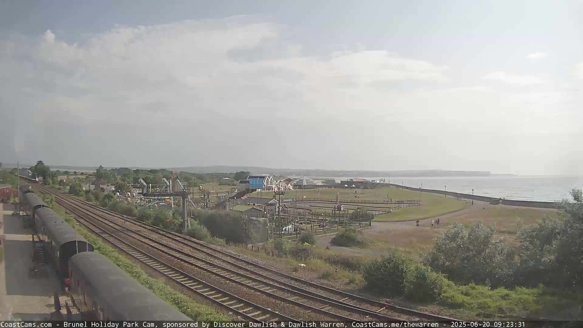 A seaside amusement park and train tracks are visible under a mostly cloudy sky, with a calm ocean in the background.