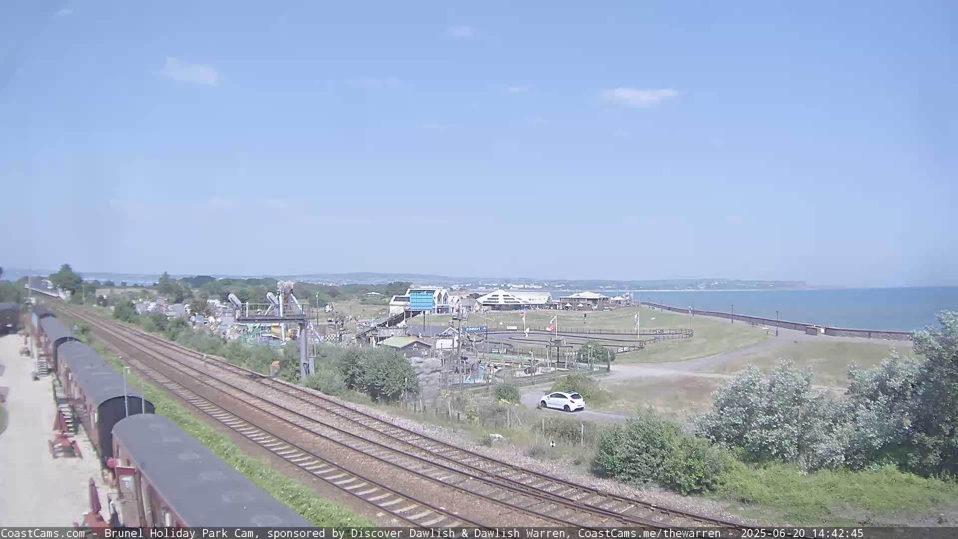 Under a clear blue sky, a seaside amusement park is visible beyond a railway line with several parked train cars, and a calm ocean stretches out in the distance.
