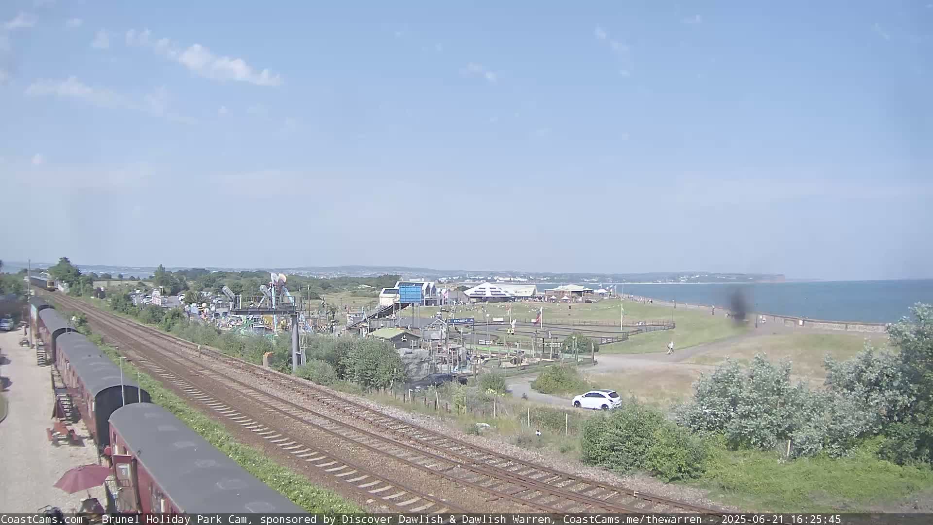 A sunny day at a seaside amusement park, featuring railway tracks in the foreground, a beach with people visible in the distance, and various park attractions.