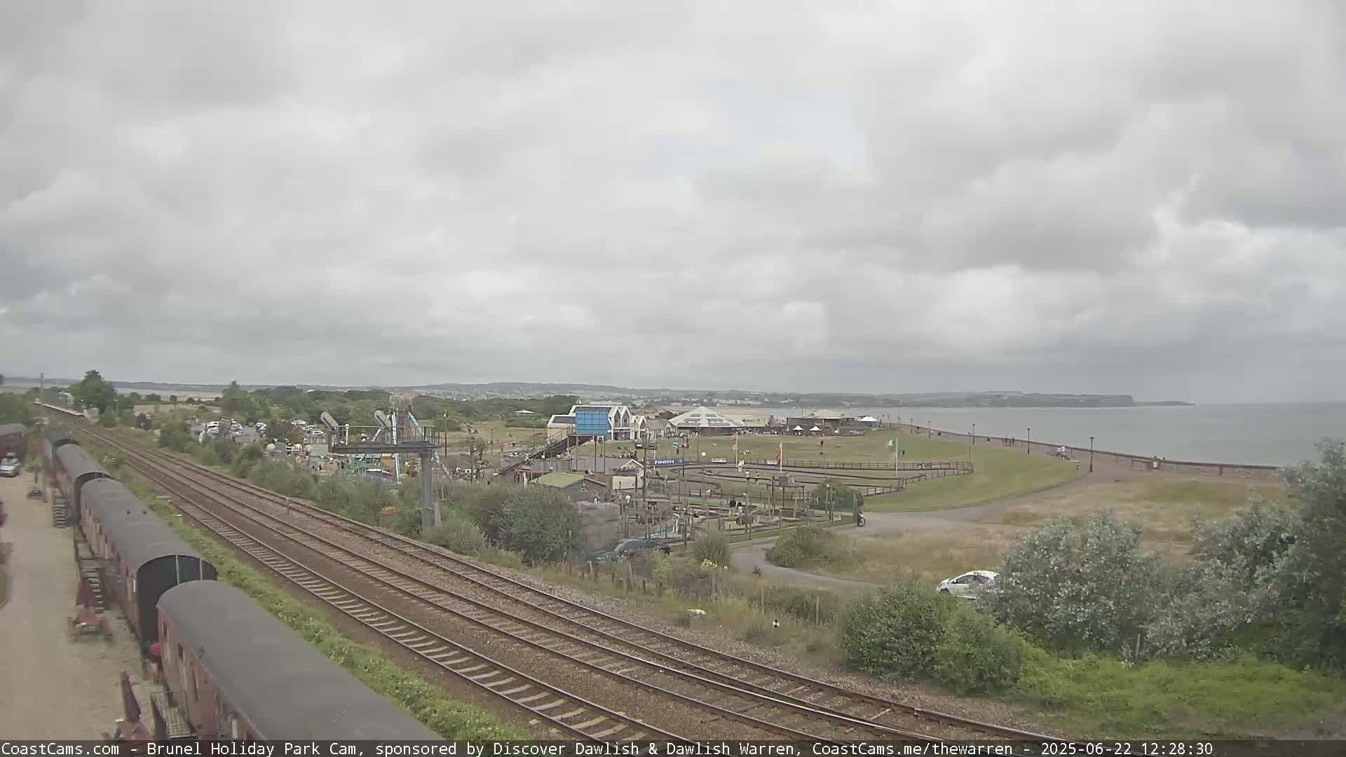 A coastal amusement park with a train track running alongside it is seen under an overcast sky, near a calm sea.
