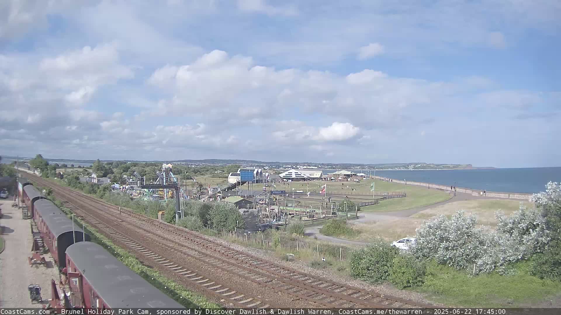A seaside amusement park with a playground and train tracks is visible under a partly cloudy sky.