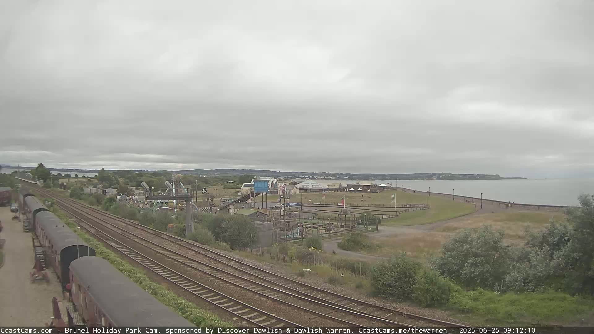 A coastal amusement park with an overcast sky and calm water is visible from a high vantage point near railroad tracks.