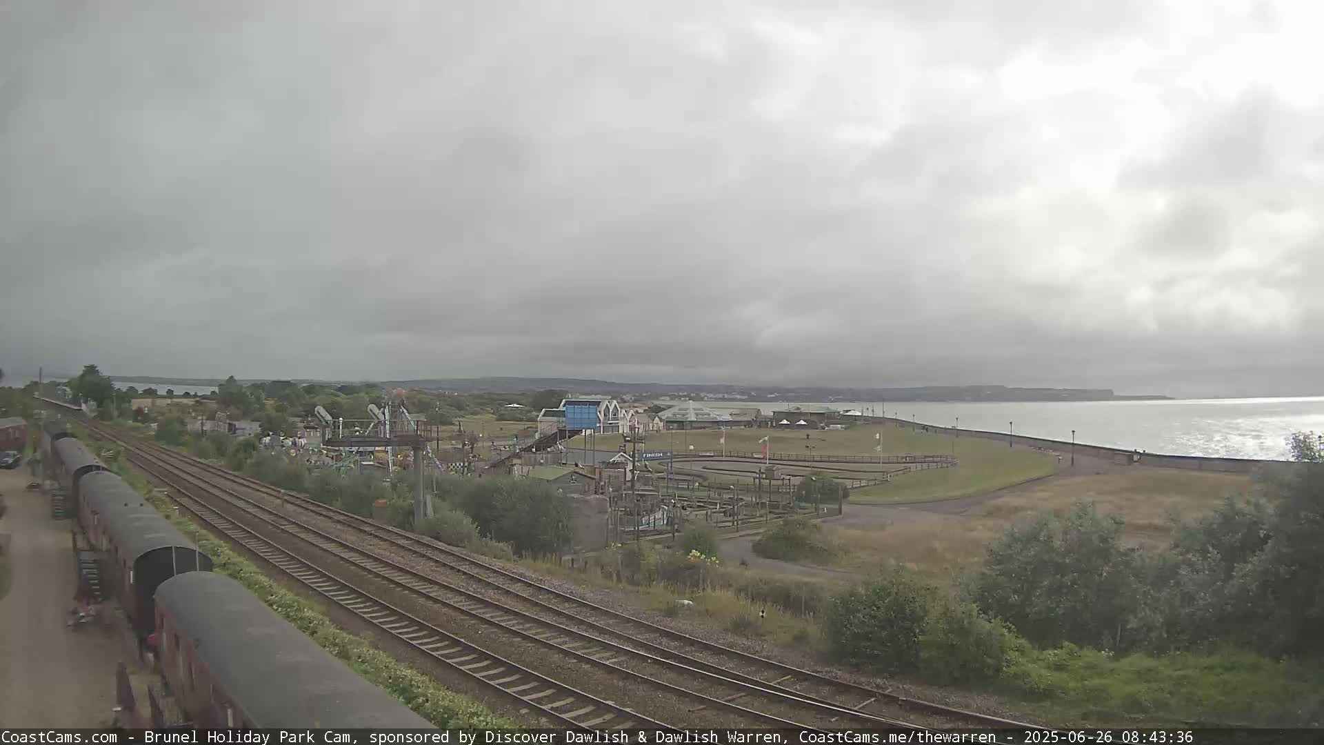 A coastal amusement park is shown under an overcast sky, with railway tracks and parked rail cars in the foreground.
