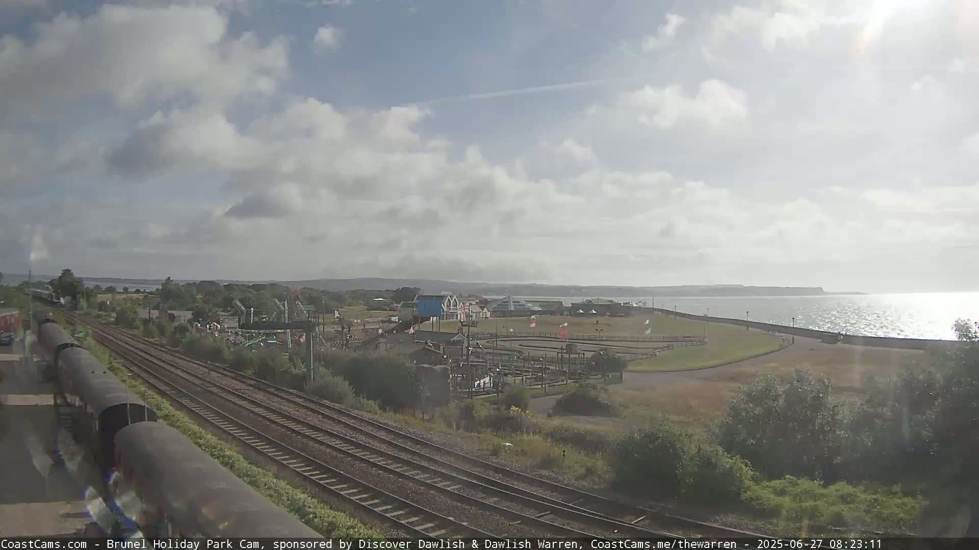 A train track runs beside a seaside amusement park under a partly cloudy sky.