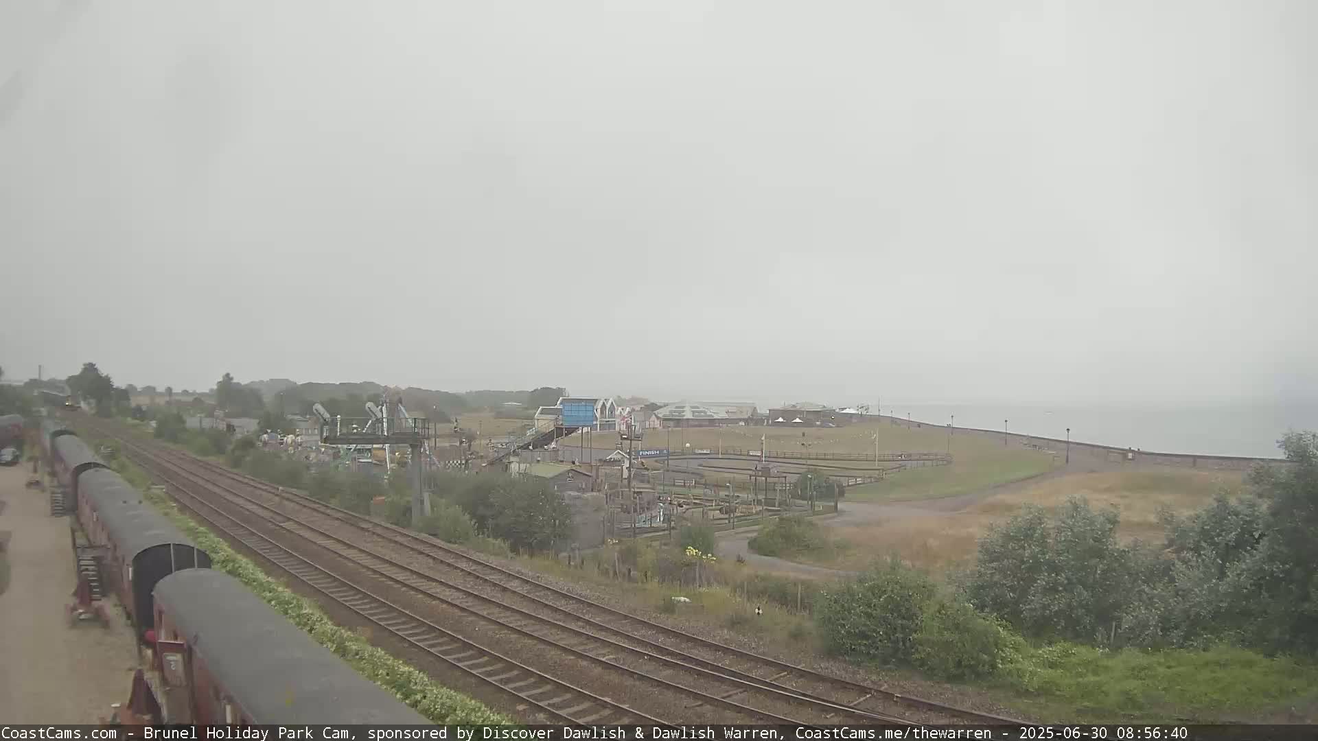 A long shot shows a train track running parallel to a seaside amusement park on an overcast day.