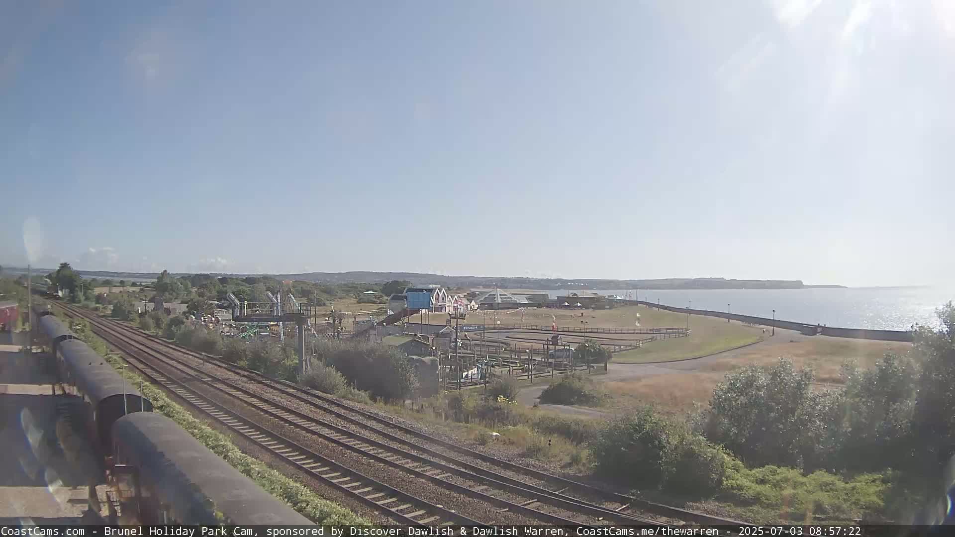 A seaside amusement park with go-karts and a playground is visible next to railroad tracks on a sunny day, with calm water in the background.
