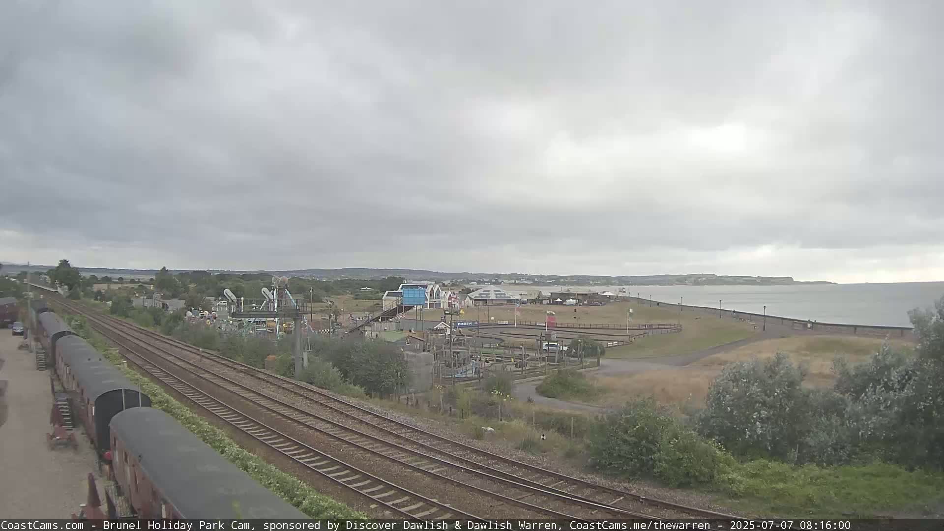 Overcast skies loom over a coastal scene featuring railway tracks, a holiday park with various structures, and a calm body of water with a shoreline.
