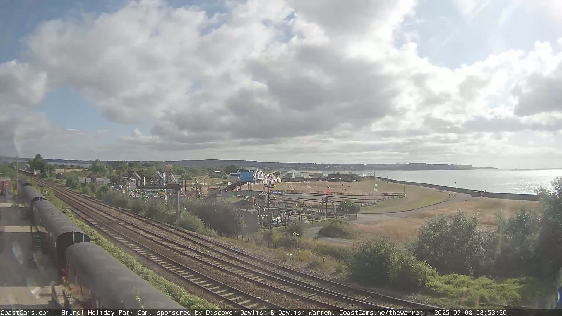 A mostly cloudy day shows a seaside amusement park next to railroad tracks and a calm body of water.