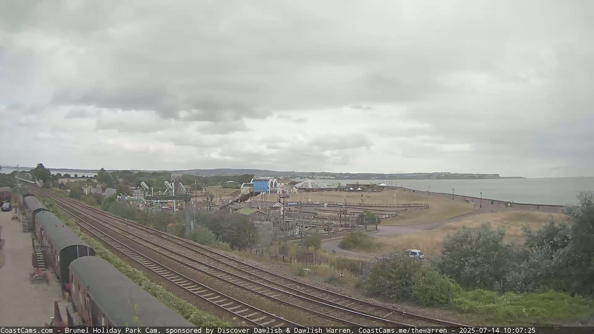 Under an overcast sky, a coastal amusement park sits adjacent to a body of water and railway tracks.
