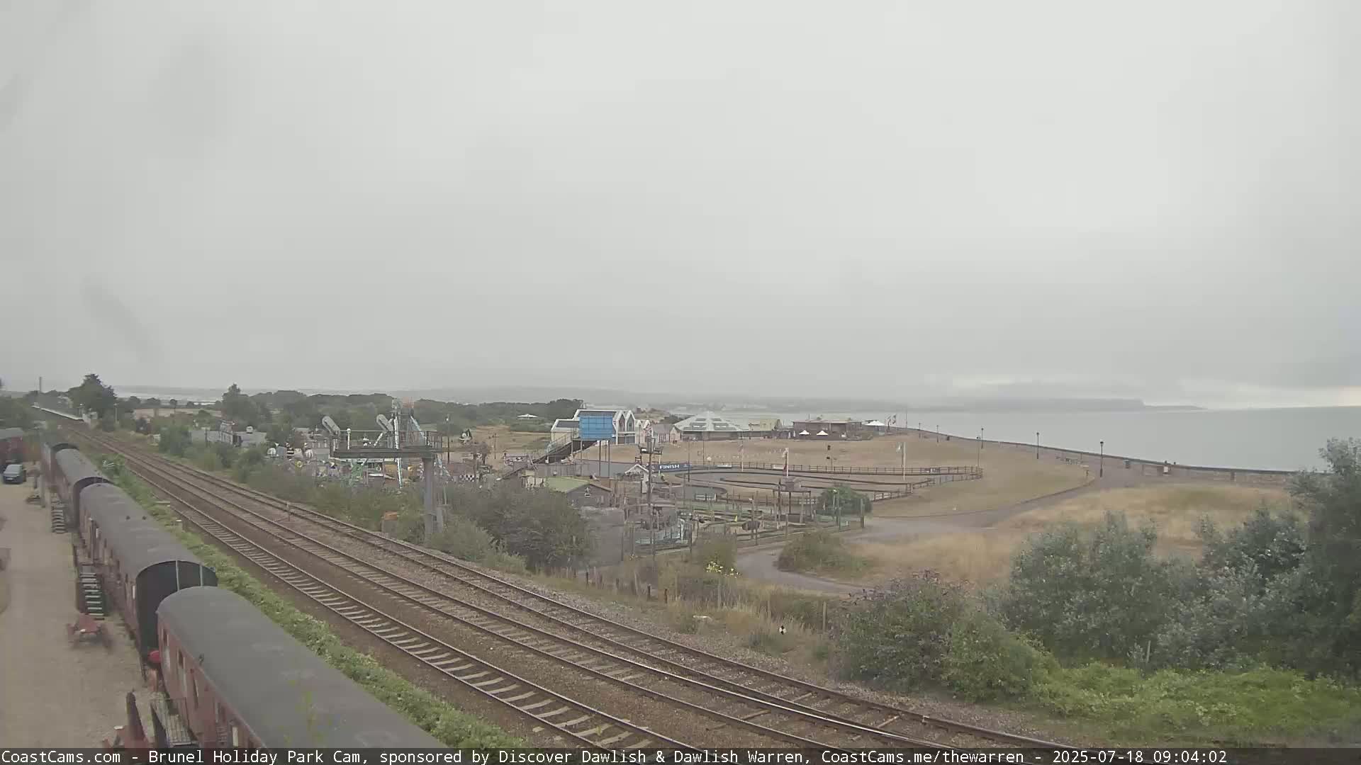 A coastal amusement park with railway tracks in the foreground is visible on an overcast day.