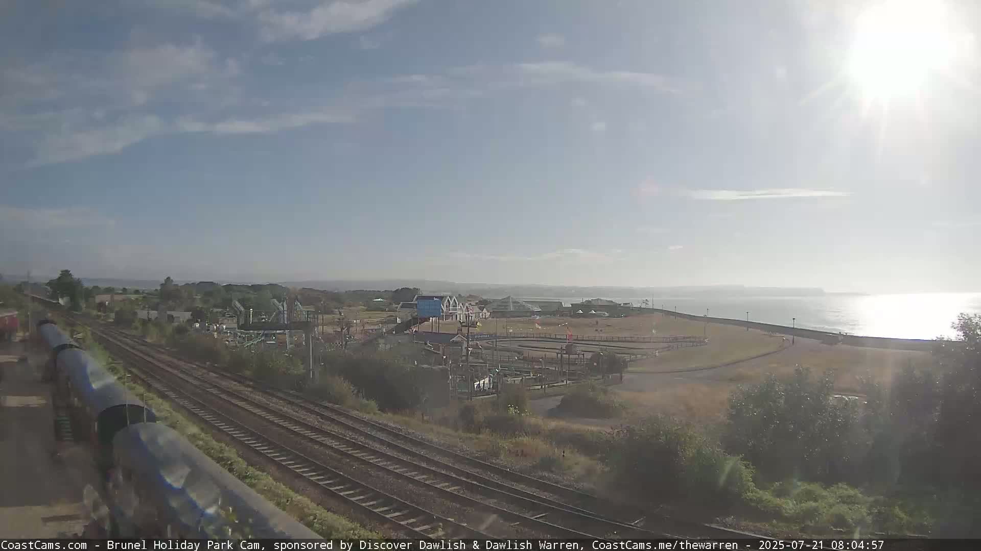 A mostly sunny day reveals a seaside amusement park near train tracks and a calm body of water.
