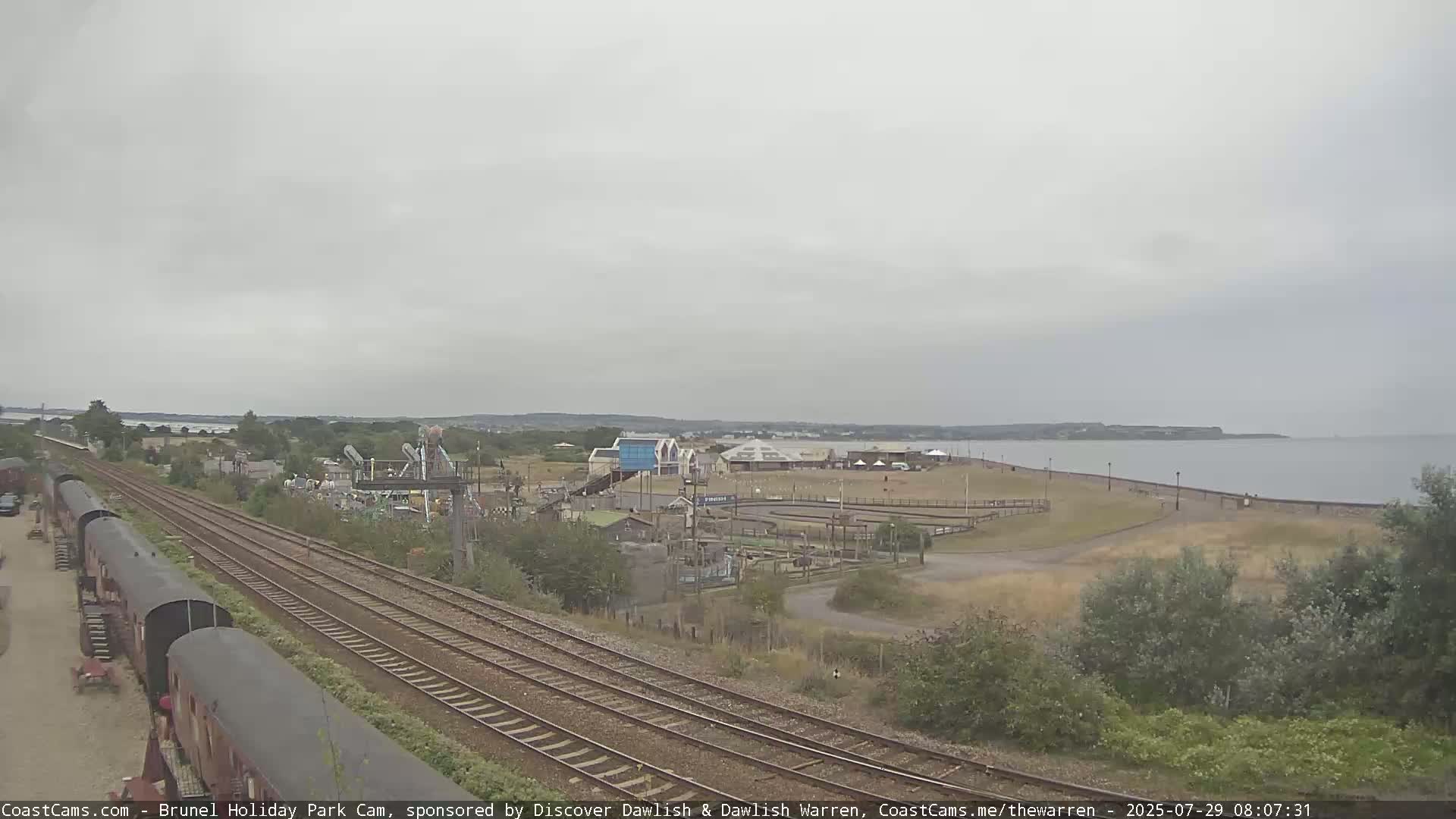 A coastal scene under an overcast sky shows railway tracks leading toward a seaside amusement park and a calm body of water.