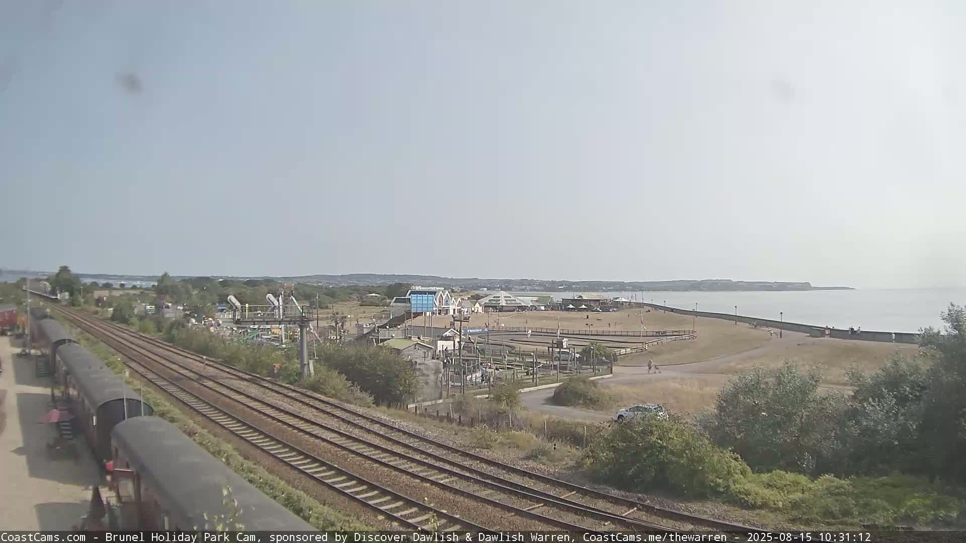 A coastal scene under an overcast sky shows railway tracks leading toward a seaside amusement park and a calm body of water.