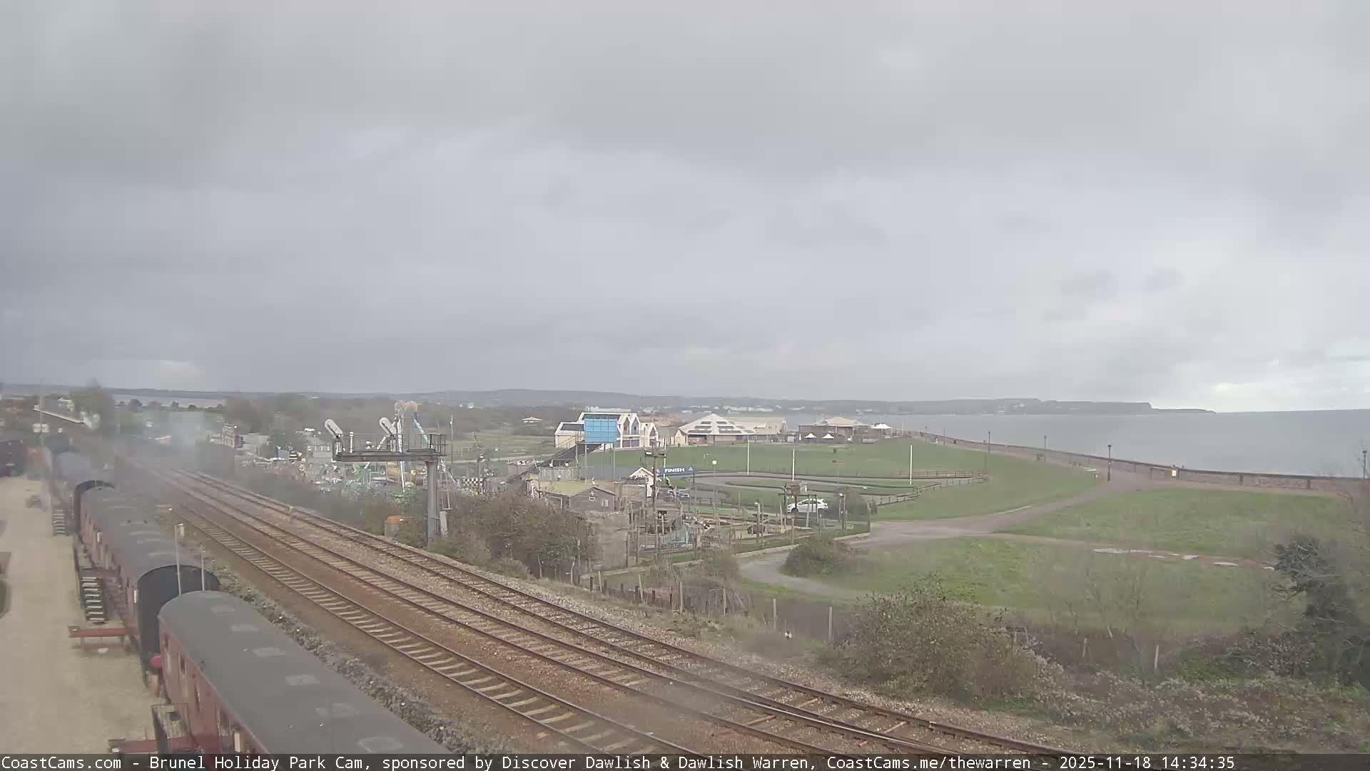 A coastal scene under an overcast sky shows railway tracks leading toward a seaside amusement park and a calm body of water.