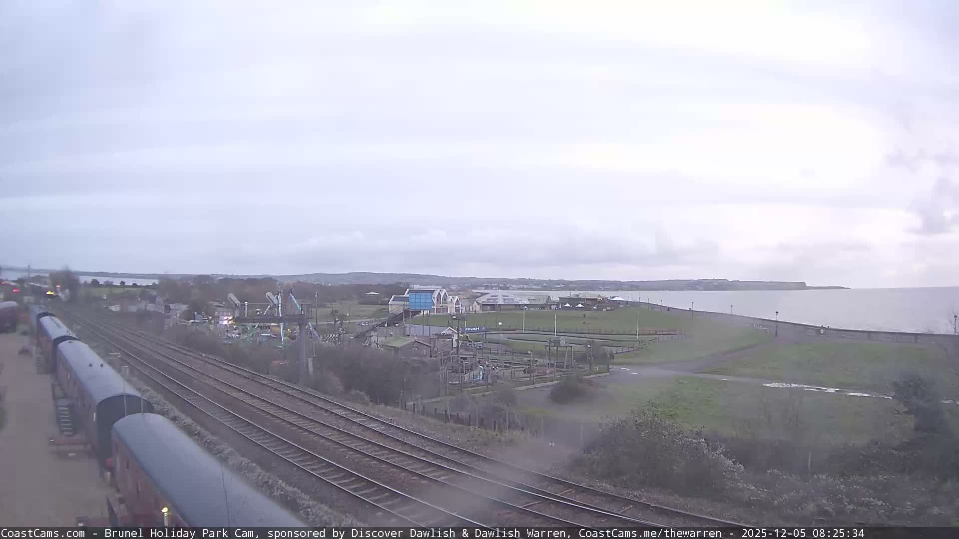 A long train sits on multiple tracks in the foreground next to a holiday park with various attractions, all under an overcast sky bordering a calm body of water with a distant coastline.