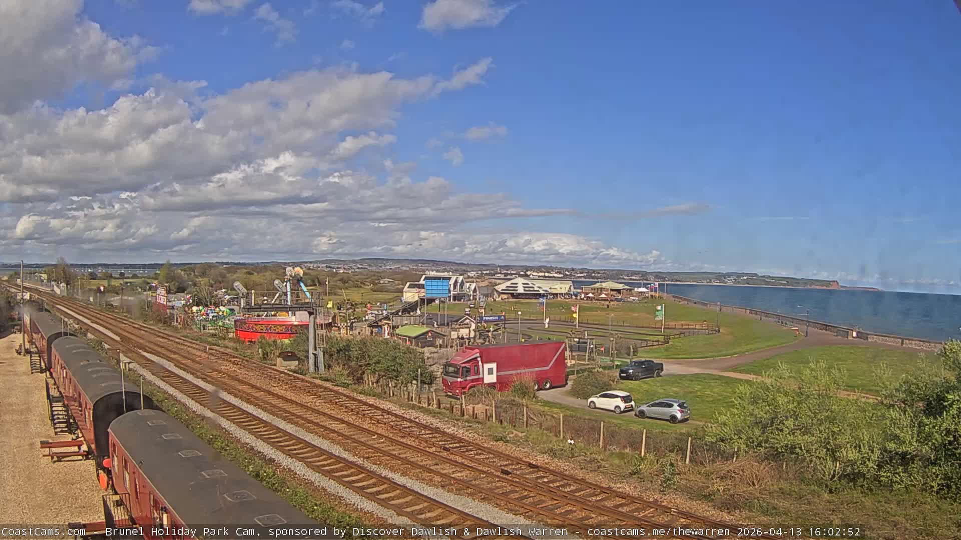 A long train sits on multiple tracks in the foreground next to a holiday park with various attractions, all under an overcast sky bordering a calm body of water with a distant coastline.