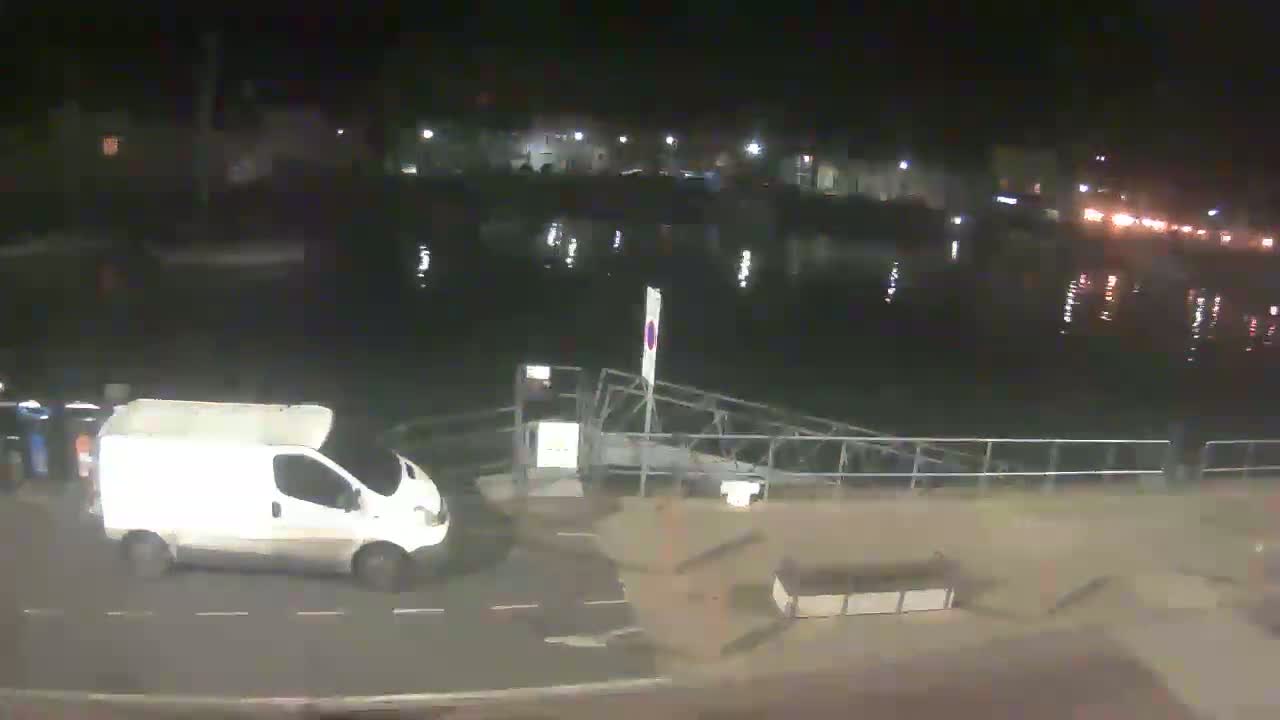 A white van is parked near a metal dock at night beside a body of water reflecting city lights.
