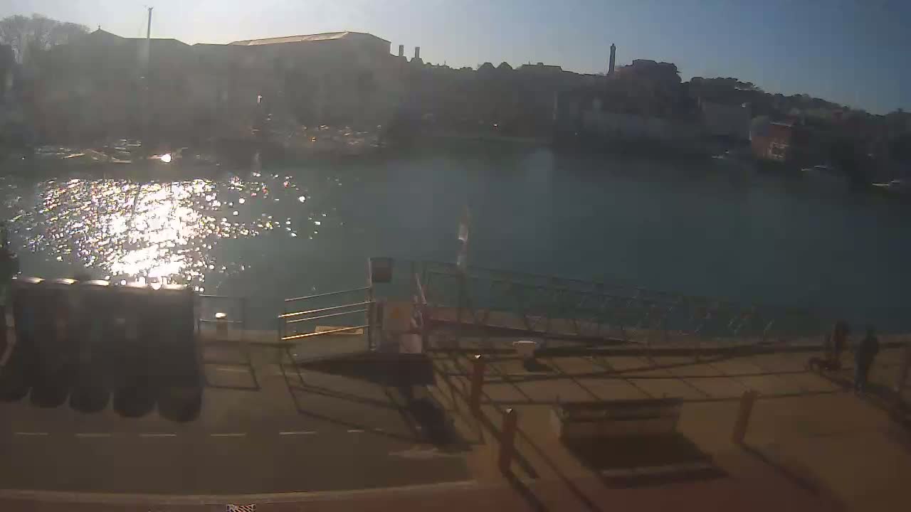A sunny day shows a waterway reflecting sunlight, with buildings and a dock in the background.