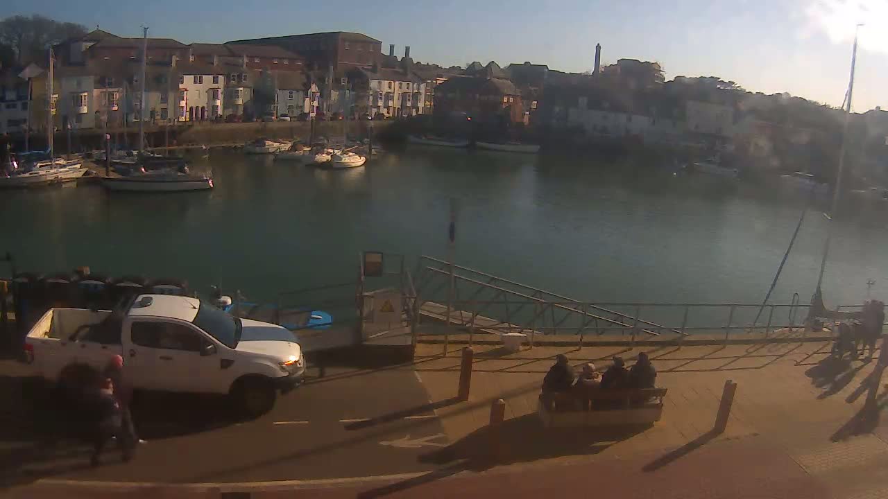 A harbor filled with boats on a sunny day, viewed from a high vantage point, showing a white pickup truck parked near a walkway with people sitting on a bench.