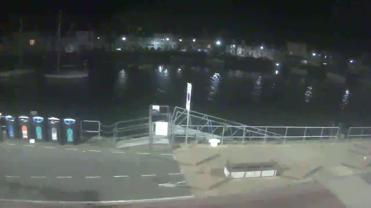 A nighttime view of a calm harbor with a boat ramp and several recycling bins, under a clear, dark sky.