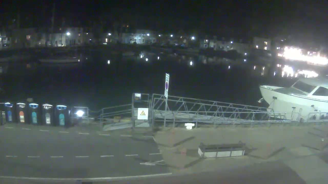 A nighttime view of a harbor with a docked boat, a metal gangway, and several recycling bins near a paved area.