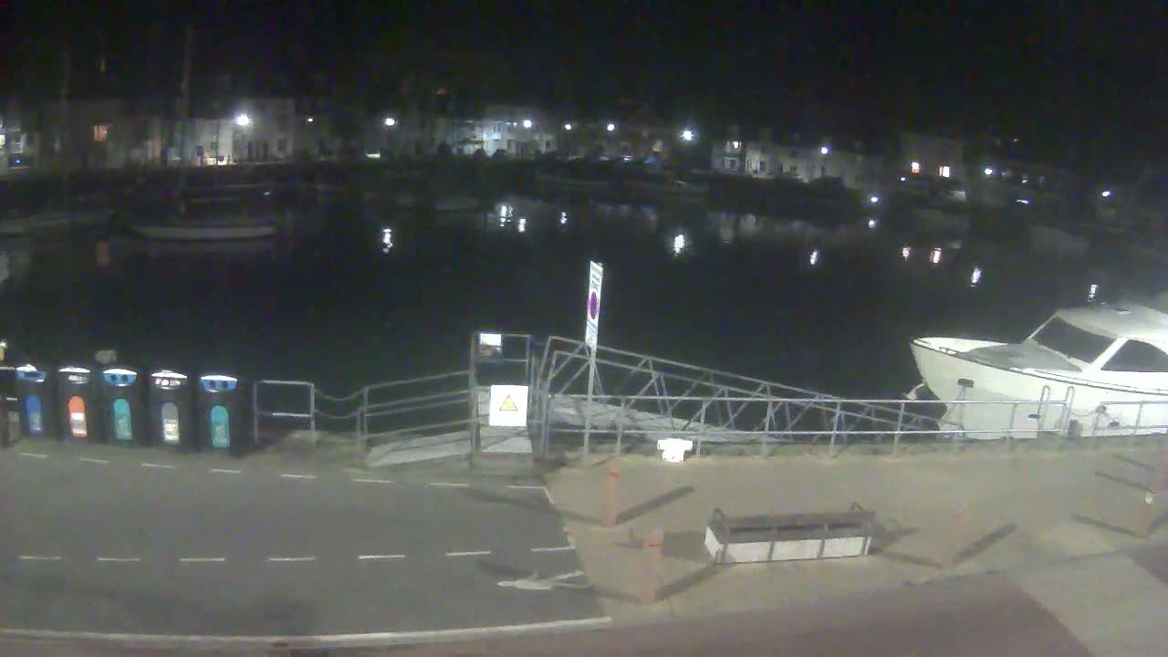 A nighttime view of a calm harbor with several boats moored, a metal gangway leading to a dock, and a row of recycling bins.