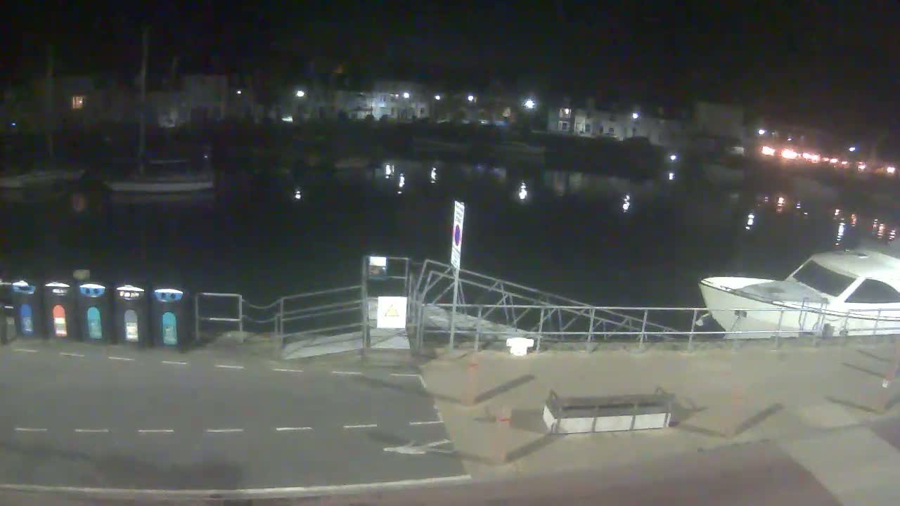 A nighttime view of a calm harbor with several boats moored, a small dock with a ramp, and a row of recycling bins on the shore.