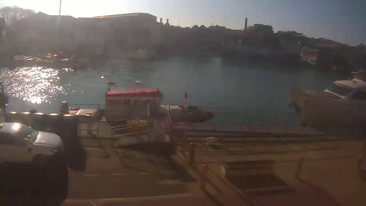 A harbor scene on a sunny day shows several boats docked near a waterfront road, with buildings and trees visible in the background.