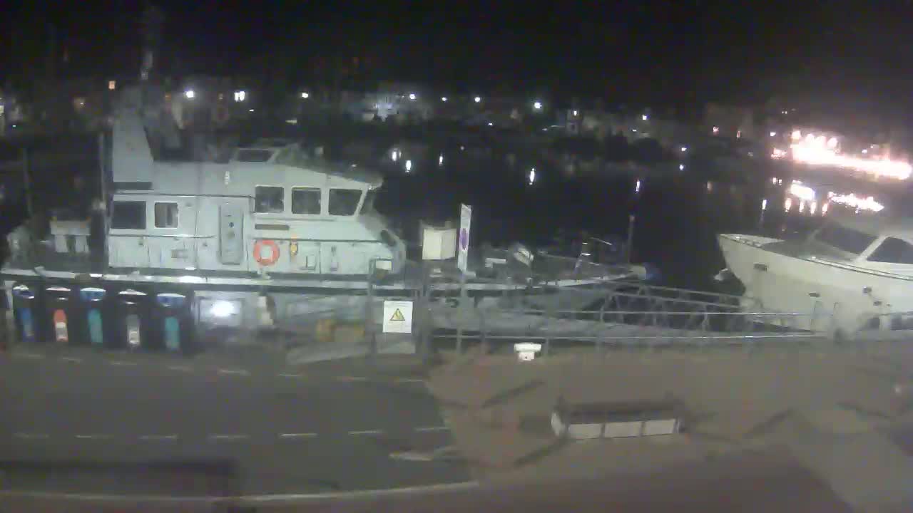 At night, a large gray patrol boat is docked next to a smaller white yacht in a harbor with lights visible in the background.