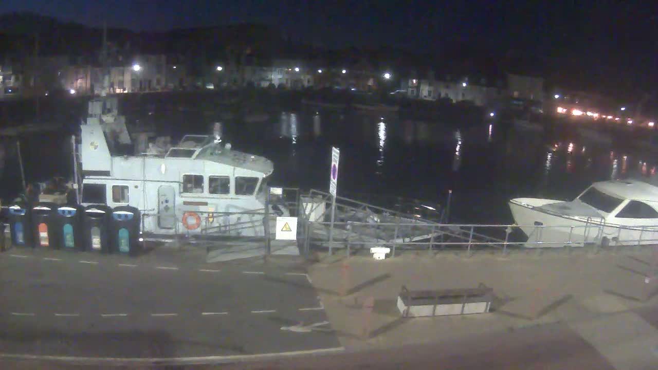 A large, light-grey boat is docked at a harbor at night, next to a smaller white motorboat, with a dark, calm body of water reflecting lights from buildings in the background.
