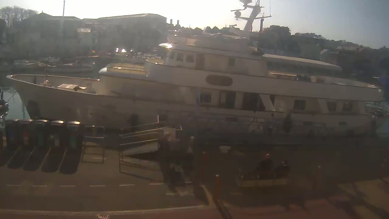 A large, light-colored yacht is docked at a harbor on a sunny day, with several smaller boats visible in the background.