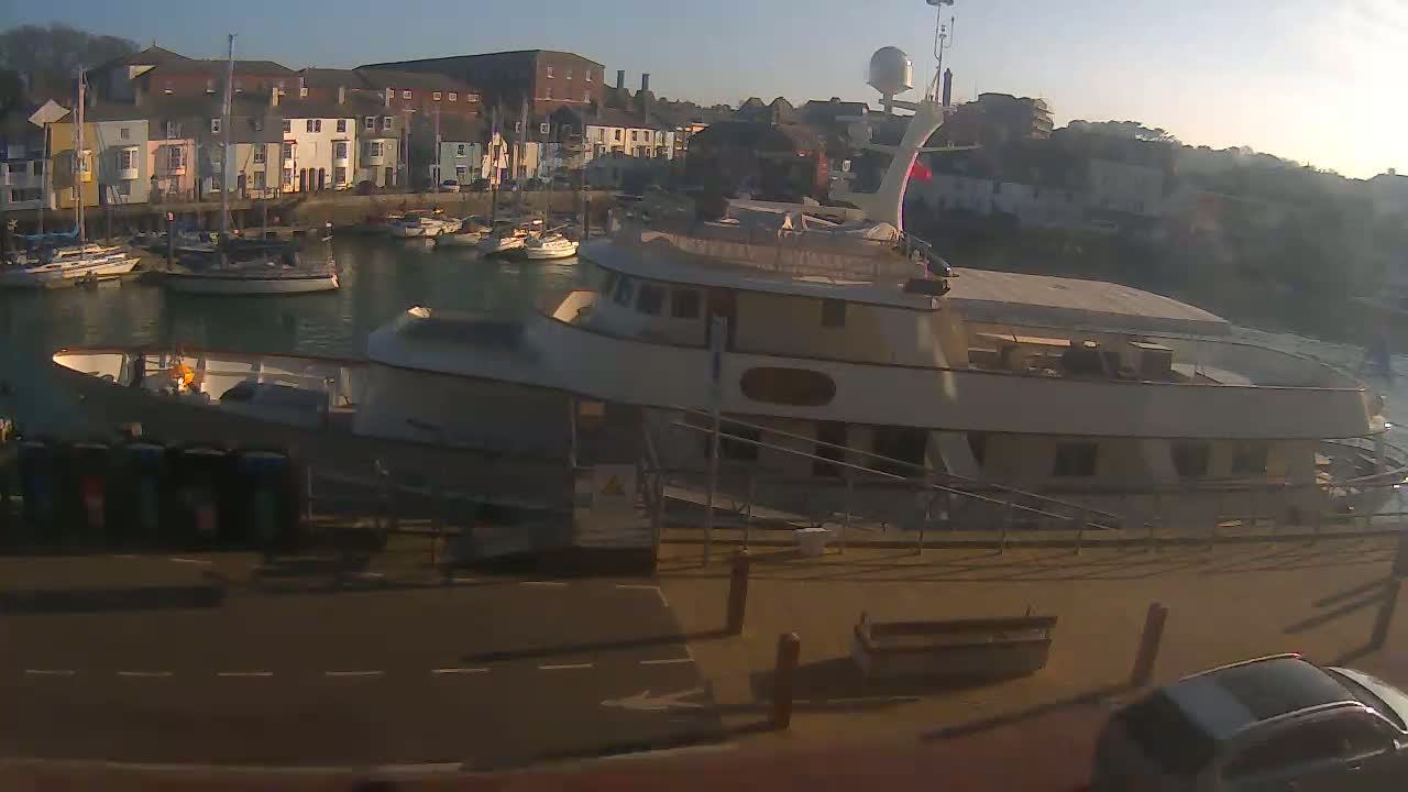 A large motor yacht is docked at a harbor filled with smaller boats, under sunny skies.