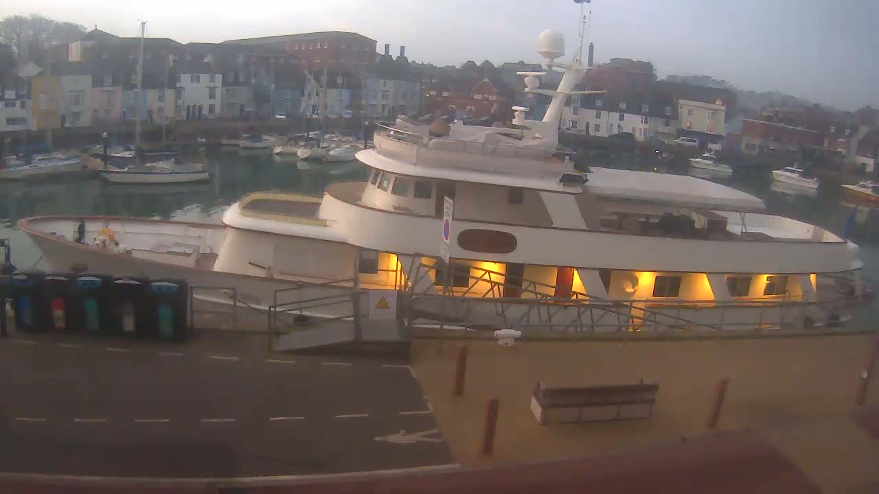 A large motor yacht is docked at a marina on an overcast day, with other boats and colorful buildings visible in the background.