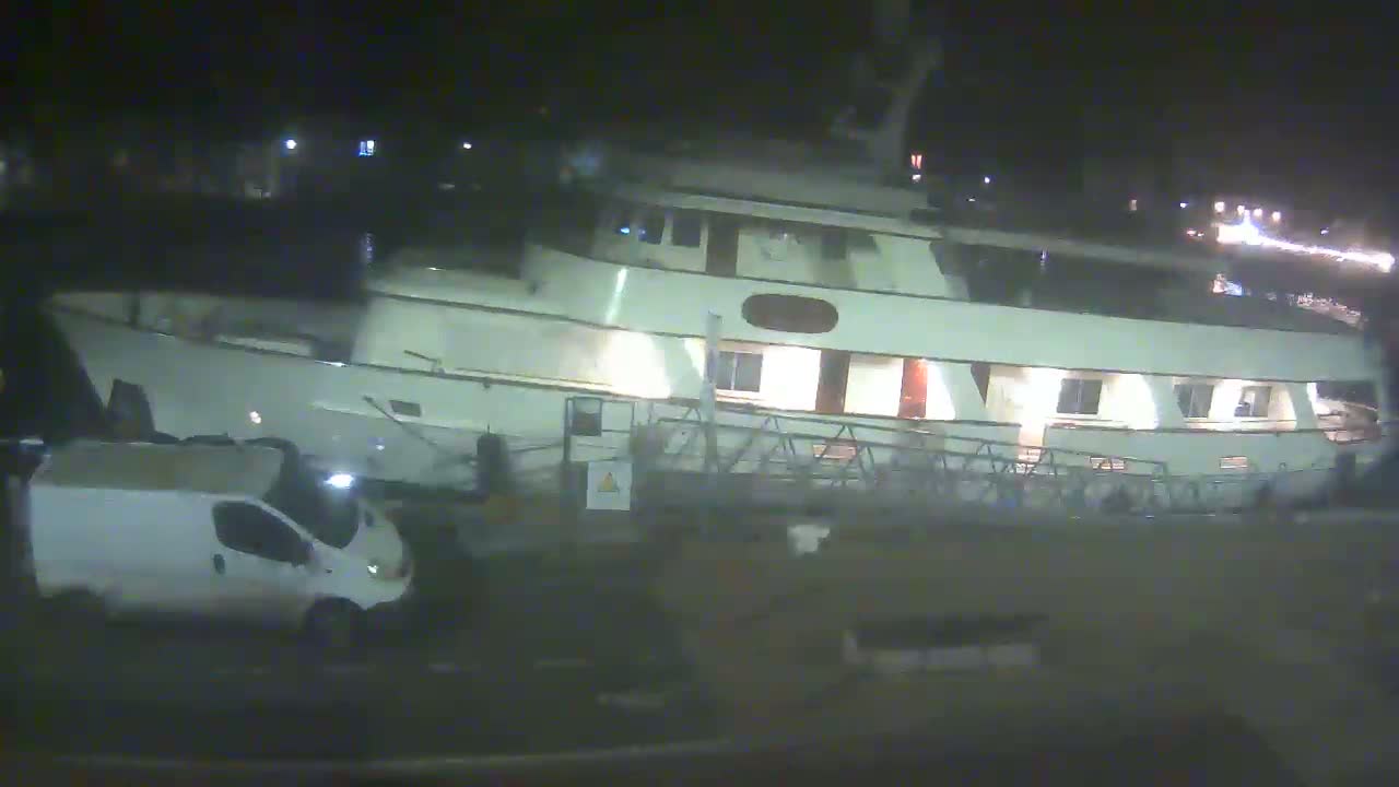 At night, a large, light-colored yacht is docked near a white van.