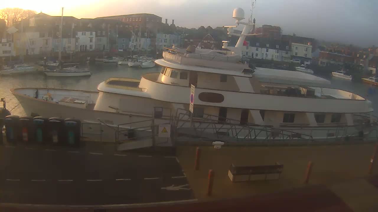 A large motor yacht is docked at a marina at sunrise, with numerous smaller boats visible in the harbor.