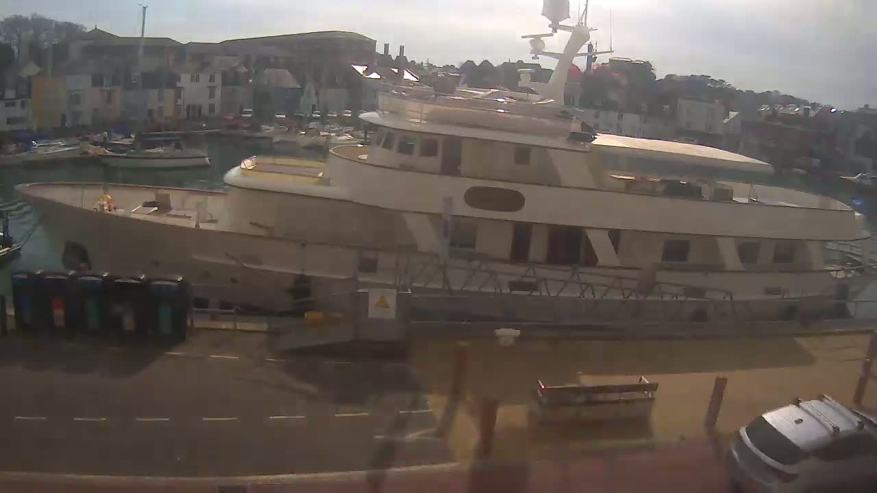 A large, light-beige motor yacht is docked in a harbor on a sunny day, with other smaller boats visible in the background.