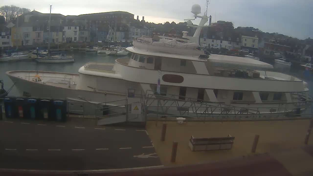 A large, white motor yacht is docked in a harbor on an overcast day, with smaller boats and buildings visible in the background.