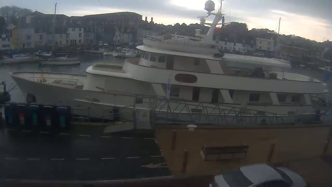 A large, light-colored yacht is docked at a harbor, with overcast skies and several smaller boats visible in the background.