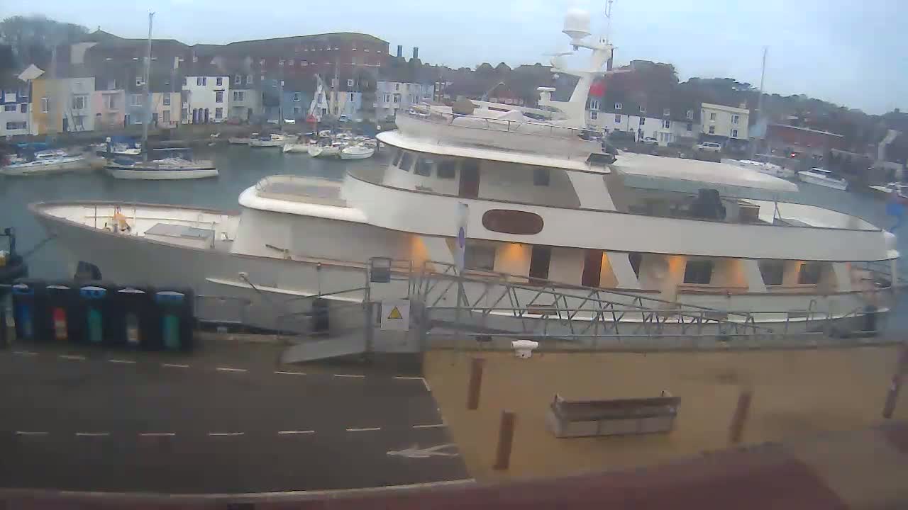 A large motor yacht is docked at a marina on an overcast day, with smaller boats visible in the background.